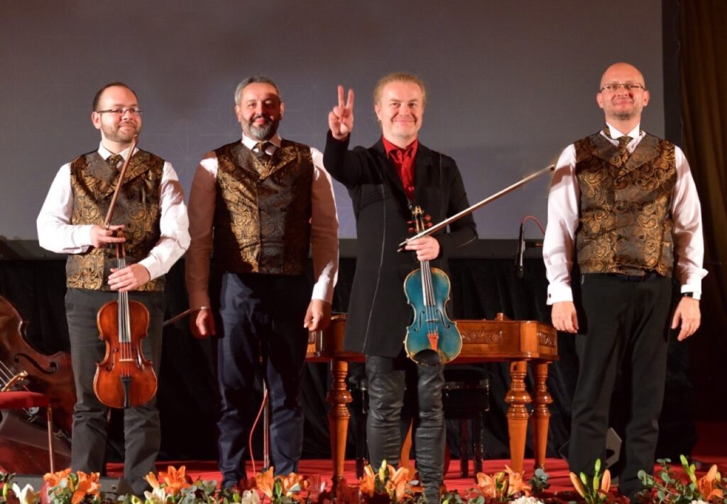 A group photo of Pavel Šporcl & Gipsy Way shows the four members, including violist Bélu Botoš (far left) and violin virtuoso Pavel Šporcl (third from left) holding his famous blue violin and greeting the audience with a V-shaped gesture.