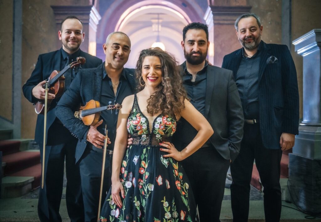 The group photo shows five people forming the Manuša group. Among them are the violist Bélu Botoš, positioned on the far left, and the singer Júlia Kozáková, standing in the middle.